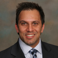 Professional headshot of a smiling man wearing a dark suit and blue striped tie, against a neutral backdrop.